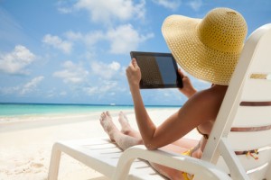 Woman holding tablet computer sitting on the beach in deck chair and taking sun bath